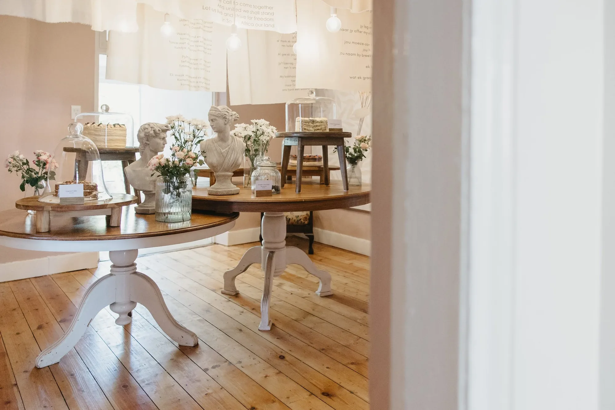 Inside the front room: cakes under glass cloches on round wooden tables, plaster busts and jars of flowers, bare bulbs strung above.